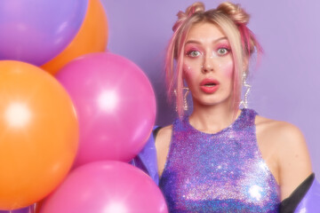 Fototapeta na wymiar Surprised woman looks shocked at camera dressed in fashionable clothes poses near inflated colorful balloons prepares for party celebrates birthday poses against purple background. Slow glow effect