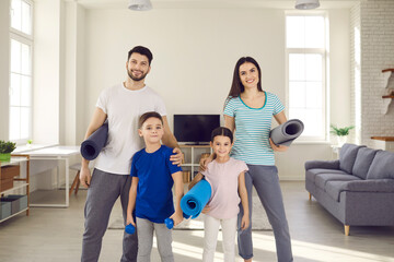Portrait of happy healthy fit young family with little children standing together, holding exercise mats and dumbbells, smiling and looking at camera. Active sports workout in lockdown at home concept