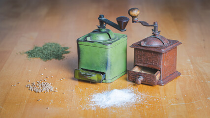 In a simple yet refined background two retro style handmade grinders from grandma&rsquo;s kitchen, one brown, one green color surrounded with cooking spices placed on a natural light brown wooden table.