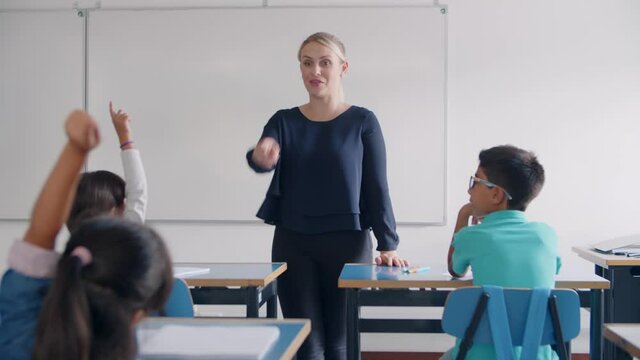 Friendly teacher giving and explaining task to pupils, asking questions in class. Students raising hands at desks. Teaching or primary education concept