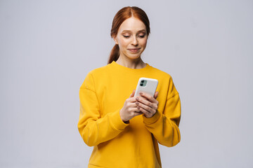 Smiling lovely young woman wearing stylish yellow sweater texting on her cell phone on isolated...