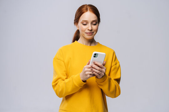 Smiling lovely young woman wearing stylish yellow sweater texting on her cell phone on isolated white background.Pretty redhead lady model emotionally showing facial expressions in studio, copy space.