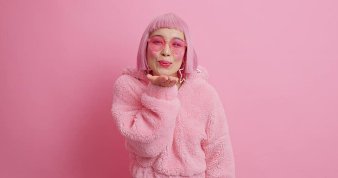 Affectionate lovely tender Asian woman has bob hairstyle blows air kiss at camera flirts with beloved person wears trendy pink sunglasses and fashionable winter coat poses indoor. Mwah to you