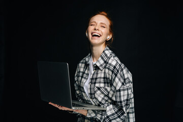 Laughing young woman wearing wireless earphones holding laptop computer and looking at camera on isolated black background. Pretty redhead lady model emotionally showing facial expressions.