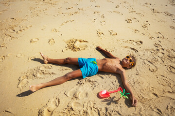 Happy little boy resting on the sand tanning on the beach near plastic toys