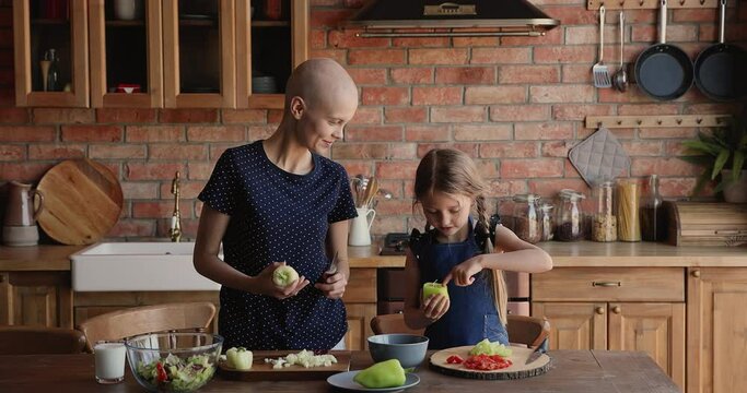 Mother cancer disease patient teaching little daughter to cook, preparing vegetable salad, cutting paprika talking standing in cozy modern kitchen. Healthy eating, time together, hobby, bond concept