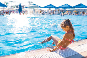 A lovely girl sits on the side of the pool with her feet dipped into clean transparent water enjoying the warm bright sun