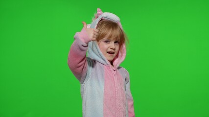 Young little blonde child 5-6 years old smiling, showing thumbs up gesture, agree sign in unicorn costume on chroma key green background. Portrait of kid girl animator in unicorn pajamas. Copy space