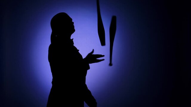 Black silhouette of a man juggling with pins in the spotlight. Circus performer with an entertaining show program. Blue backdrop illuminated by studio light. Close up. Slow motion.