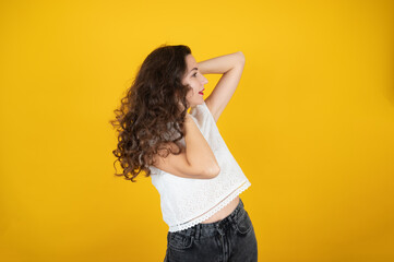 Stylish beautiful caucasian woman in a blouse and jeans standing on yellow background. Young woman touches her curly hair with her hands, side view