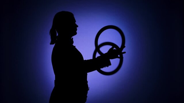 Side view black silhouette of male juggler rotating rings in the spotlight. Professional circus performer demonstrates his abilities. Blue backdrop illuminated by studio light. Close up. Slow motion.