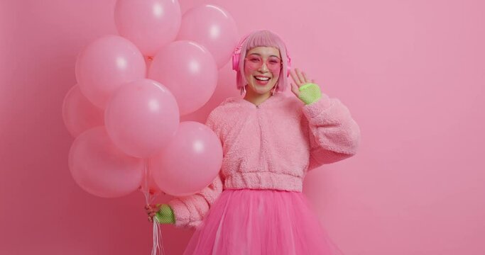 Joyful pretty woman with pink bob hair waves hello greets friends on party wears stereo headphones winter coat and pleated skirt holds inflated balloons celebrates something expresses happiness