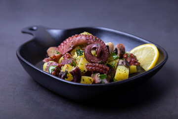 Warm salad with octopus, potatoes and lemon in a black small pan. Close-up, black background.