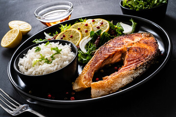 Fried salmon steak with basmati rice and  mix of vegetables served on black plate on wooden table
