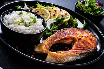Fried salmon steak with basmati rice and  mix of vegetables served on black plate on wooden table
