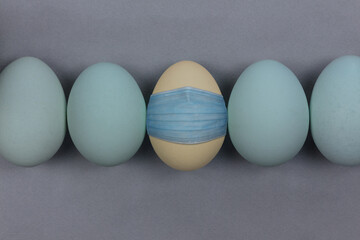  a yellow egg wearing a blue face mask in a row of blue eggs on a gray background, theme for covid or coronavirus Easter or other humorous concept, with copy space
