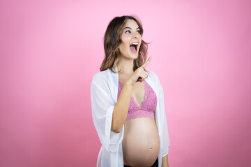 Young beautiful brunette woman pregnant expecting baby over isolated pink background smiling and pointing with hand and finger to the side