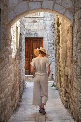 Fototapeta na wymiar Rear view of beautiful blonde young female traveler wearing straw sun hat sightseeing and enjoying summer vacation in an old traditional costal town at Adriatic cost, Croatia.