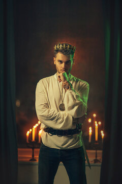 Portrait of handsome attractive man in image of medieval king. Vintage clothing historic white shirt retro clothing gold crown on head. Dark gothic room, studio. Fashion model. Adult sexy guy knight