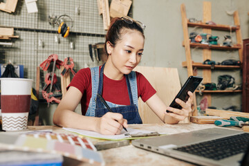 Young serious Vietnamse female carpenter sitting at desk, reading text messages on smartphone and writing in planner