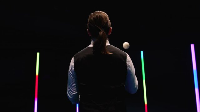 Orbital shot professional circus performer juggling with five white balls against a black studio background amid bright neon lights. Close up. Slow motion.