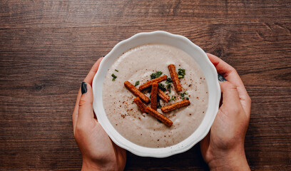 Woman hands holding bowl of Mushroom cream soup with croutons, herbs and spices on a wooden background. with copy space. top view