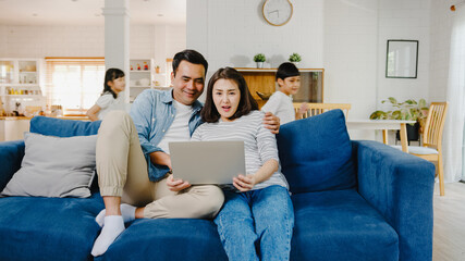 Asia family dad and mom sit on couch enjoy shopping online on laptop while daughter and son have fun shouting run around sofa in living room at home. Social distancing, Quarantine for coronavirus.