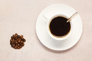 A cup of freshly brewed coffee cup and spoon having some roasted coffee beans laying beside