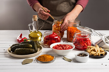 The concept of fermented canned vegetarian food. Carrots, tomatoes, cucumbers, pickled, sauerkraut sour in glass jars and plates on a white wooden kitchen table. Home-made preparations.