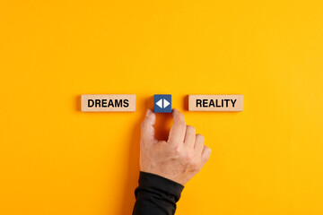 Male hand holds a wooden cube with arrow icon between the options of dreams or reality.