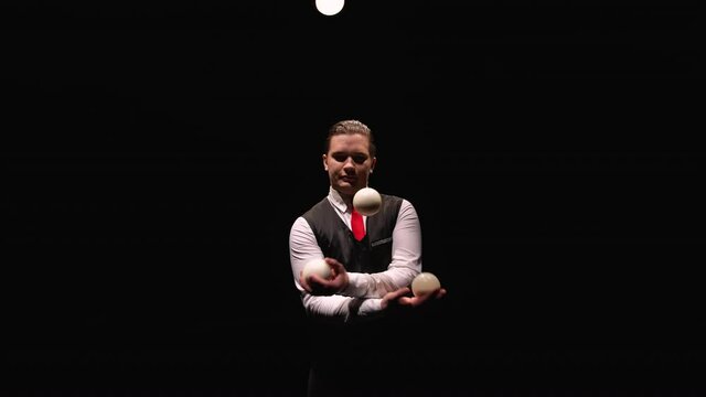 Orbital shot of a stylish man professionally juggling white balls. Circus performer illuminated by lights throws and catches balls on black studio background. Close up. Slow motion.