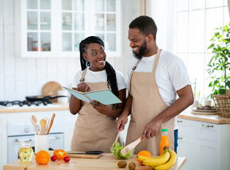 Happy Black Couple Checking Recipe In Cookbook While Cooking Together In Kitchen