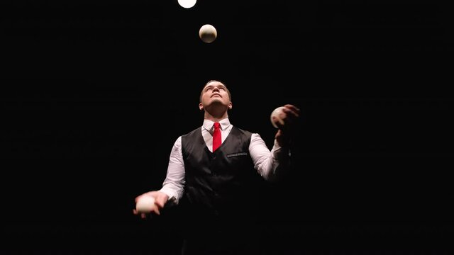Orbital shot of a stylish man professionally juggling white balls. Circus performer illuminated by lights throws and catches balls on black studio background. Close up. Slow motion.