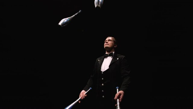 Camera rotates around circus juggler using pins to juggle. A man in a black suit performs exciting tricks in a dark backlit studio. Orbital shot close up. Slow motion.