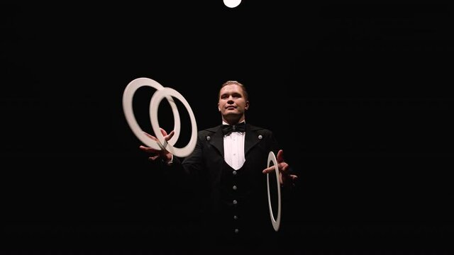 Camera rotates around showman, trowels twisting white rings. Juggler, illuminated by lights, shows tricks on a black studio background. Orbital shot close up. Slow motion.