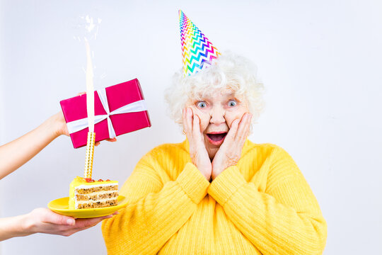 Older Woman With A Gift Wear Yellow Sweater And Horn Cap On A White Background Holding Plate With Cake With Fireworks