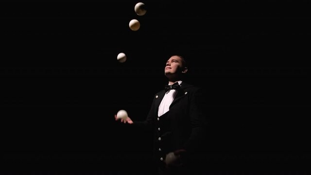 Orbital shot of a man in a black suit professionally juggling white balls. A circus performer, illuminated by lights, throws and catches balls in a dark studio. Close up. Slow motion.