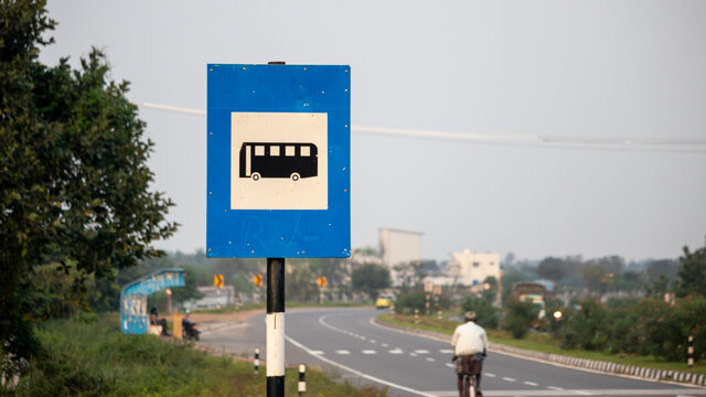 Clear wide angle photo of Indian National Highways Bus Stop Road Sign Board with Blue white background and Bus Logo in black colour in middle with road and bus stop in background where people get on