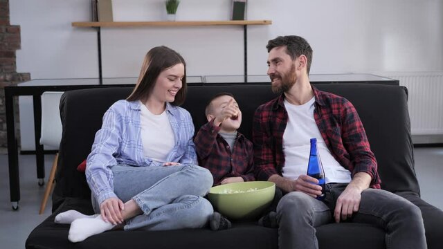 Close up of young family of three people watching TV laughing feeling happy together. Husband wife and little son spending leisure time watching movie eating popcorn. Parents and children.