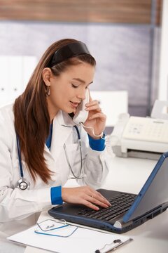 Young female doctor sitting at desk in doctors office using computer, working.