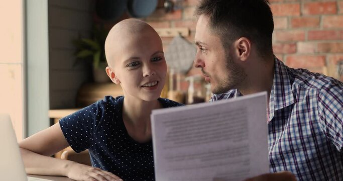 Bald young woman cancer patient her loving husband sit in kitchen read test health check up analysis result hugging celebrate victory over oncology disease feel happy. Full recovery, remission concept