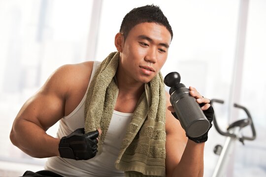 Muscular asian man resting in gym after exercise, drinking from water bottle.