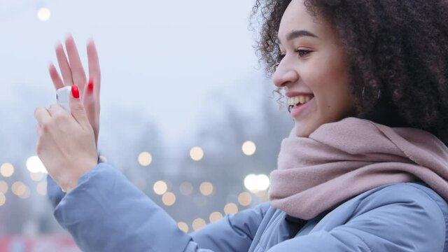 Slow motion side view friendly afro american student millennial girl wears blue jacket and pink scarf standing alone outdoors in winter holding mobile siartphone talking on video waving hand greeting