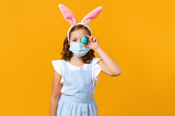 Little girl in the ears of an Easter bunny in a protective medical mask on a yellow background. The...