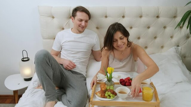 Young woman is lying in bed with the breakfast while her husband come and kiss her, happy smile, honeymoon concept.