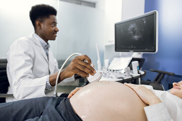 Young professional male African doctor, looking at the screen, while performing ultrasound scan of the abdomen of pregnant Caucasian woman in clinic. Focus on ultrasound transducer