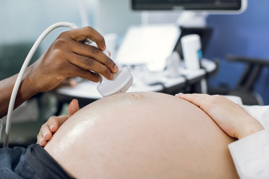 Close up cropped image of hand of male African doctor obstetrician sonographer with transducer, making ultrasound scan of pregnant belly of the female patient