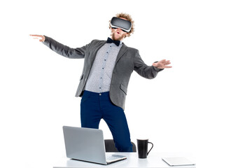 Portrait of curly-haired businessman using vr glasses while standing on chair at table with gadgets and cup of coffee.