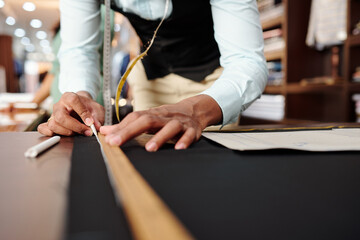 Close-up image of tailor using wooden ruler and piece of chalk when drawing detail of jacket on dark wool fabric