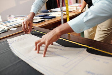 Hands of tailor tracing sewing pattern on black fabric when making trousers for client